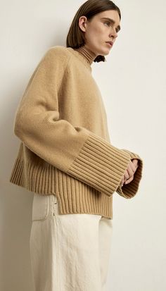 a woman standing in front of a white wall wearing a tan sweater and cream pants