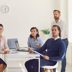 three people sitting at a table with laptops and papers in front of them, one person standing up