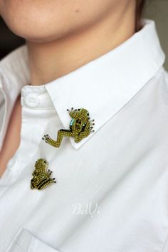 a close up of a person wearing a white shirt and two green frog brooches