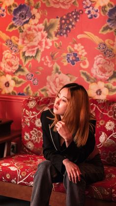 a woman sitting on top of a couch next to a red flowered wallpaper