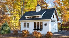 a small white house with a black roof in the fall leaves and trees around it