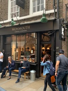 people are walking past a coffee shop on the sidewalk in front of brick buildings with green lamps