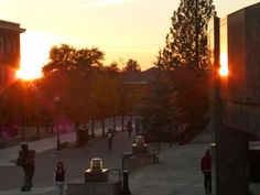 the sun is setting over some buildings and people are walking on the sidewalk in front of them