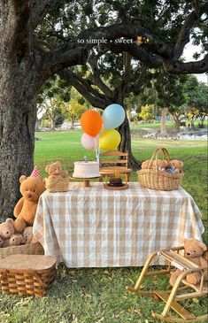 a picnic table with balloons and teddy bears in the grass under a large oak tree