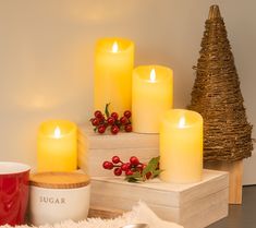 lit candles on display in front of christmas tree and mugs with holly berries next to them