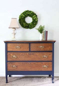 a dresser with a wreath on top and a lamp next to it in front of a white wall