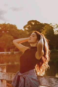 a woman sitting on top of a wooden bench next to a lake with trees in the background