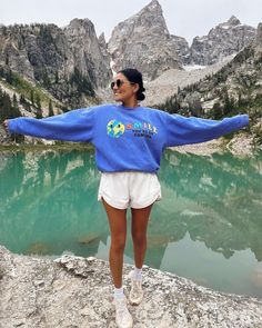 a woman standing on top of a rock next to a lake with mountains in the background