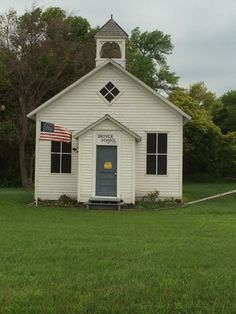 a small white church with an american flag on the front door and side window, in a grassy field