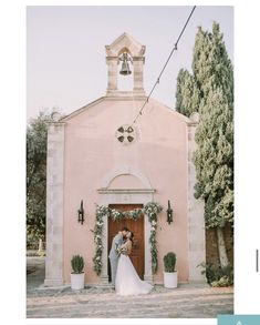 a bride and groom standing in front of a pink church with potted plants on either side