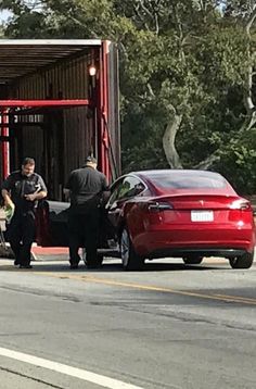 two men are standing next to the back of a red car