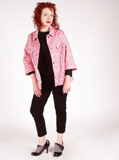 a woman with red hair wearing black pants and a pink floral print jacket stands in front of a white background