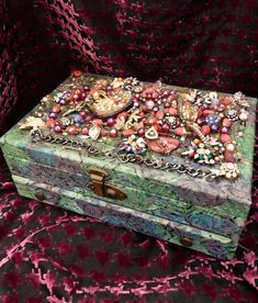 an ornate box is covered with beads and other jewelry on a pink velvet cloth background