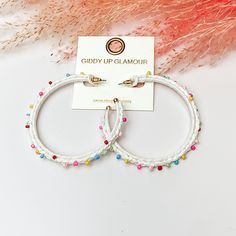 Pictured are raffia braided hoop earrings in white with colorful beads. They are pictured with a pink feather on a white background. Trendy White Beaded Earrings With Colorful Beads, Trendy White Earrings With Colorful Beads, Handmade Trendy White Beaded Earrings, White Hoop Earrings For Spring Party, White Handmade Trendy Beaded Earrings, Handmade White Beaded Earrings Trendy Style, White Spring Party Hoop Earrings, Trendy White Beaded Earrings For Summer, White Small Hoop Earrings For Spring