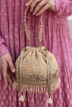 "If you want to switch up your festive style, the Monroe collection sets it apart from the other festive potli. It has a vintage appeal and will add a classic quotient to your outfit. Adorned with ethereal mirror work embroidery throughout the purse, embellished scallop lace handle, and crystals, this style is sure to make a story. Stunning mirrorwork embroidered potli that you can flaunt effortlessly. Color: Beige Height: 7\"  Base: 7.5\" x 4.5\" Handle drop: 5.5\" Material : Embroidered poly silk, poly satin lining, pearl and crystal fringe lace, woven gold thread drawstrings, embellished scallop lace handle and pearl  tassels." Bollywood Style Handwork Bags For Reception, Bollywood Style Bags With Handwork For Reception, Gold Zari Work Wedding Clutch, Bollywood Style Gold Festive Shoulder Bag, Bollywood Style Gold Bag For Reception, Traditional Gota Work Bag For Reception, Zari Work Potli Bag For Festivals And Reception, Bollywood Style Potli Bag With Dori Work For Reception, Gold Clutch With Zari Work For Wedding