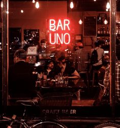 people sitting at tables in front of a bar with neon signs on the wall behind them