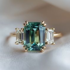 an emerald and diamond ring with three baguets on the side, sitting on a white cloth