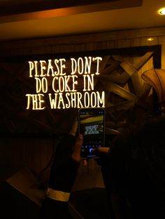 a person holding up a cell phone in front of a neon sign that reads please don't do coke in the washroom