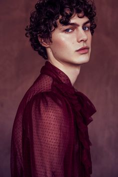 a young man with curly hair and blue eyes wearing a red shirt is looking at the camera