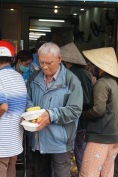 an older man holding a bowl of fruit while standing next to other people in front of a building