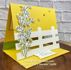 a card with flowers on it and a white fence in the background, sitting on a wooden table