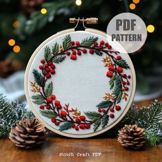 an embroidered christmas wreath with red berries, pine cones and evergreen needles on a wooden table