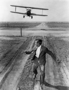 a man in suit and tie running towards an airplane flying over the dirt road that he is driving on