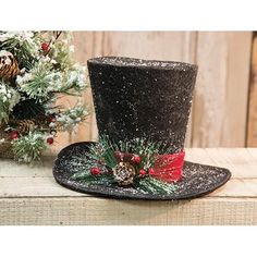 a black top hat with red ribbon and pine cones on the side, sitting next to a christmas tree