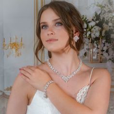 a woman in a white dress is wearing a necklace and bracelet with flowers on the wall behind her