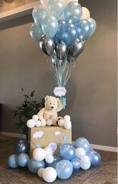 a teddy bear sitting on top of a box surrounded by blue, white and silver balloons