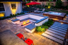 an outdoor fire pit surrounded by landscaping and patio furniture at night with red umbrellas