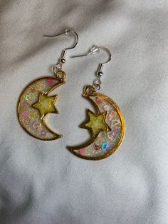 Shine like the night sky with these celestial-themed earrings. The moon and star are meticulously crafted in gold bezel settings. The moon is filled with white and clear glitter UV resin, further adorned with a heart and star-shaped confetti glitter that adds a playful touch. On the other hand, the star boasts neon yellow glitter and a glow-in-the-dark star, casting a unique glow that will certainly turn heads. Magical Gold Jewelry For Party, Celestial Star-shaped Sparkling Earrings, Gold Space-themed Earrings Gift, Sparkling Star-shaped Celestial Earrings, Space-themed Gold Earrings Gift, Celestial Star Charm Round Earrings, Crescent-shaped Earrings With Star Charm As Gift, Crescent Earrings With Star Charm As Gift, White Moon Charm Jewelry For Party