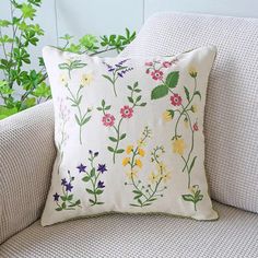 a white pillow with colorful flowers on it sitting on a couch next to a plant