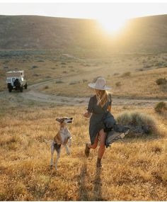 a woman in a hat and dress running with a dog on a grassy field at sunset