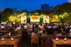 people are sitting at tables and eating in an outdoor dining area with lights on the trees