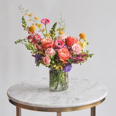 A glass vase with a medium flower arrangement sits on a marble table in front of a white backdrop. The flowers are many colors and include roses Multicolor Floral Arrangement, Bridal Shower Color Schemes Summer, Flower Arrangements For Table Birthday, Artificial Wildflower Arrangements, Live Flower Wedding Bouquets, Wildflower Bouquet In Vase, Bouquet Of Flowers Colorful, March Floral Arrangements, Colorful Summer Wedding Flowers