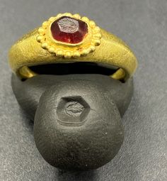 a gold ring with a red stone in the center on a gray surface next to a rock