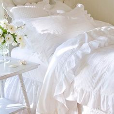 a white bed sitting next to a table with a vase on it and flowers in the corner