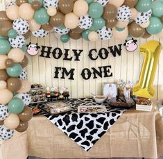 a table topped with lots of balloons and desserts next to a cow themed backdrop