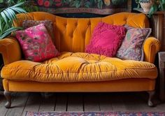 an orange couch with colorful pillows on it in front of a painting and potted plants