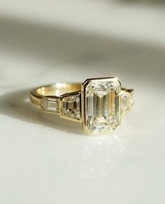 an emerald cut diamond ring with three baguets on the side, sitting on a white surface