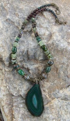 This attractive necklace is composed of many types of stones and beads:  there are 4 large tibetan line agate beads; 4 large green oval jasper gemstone beads; prehnite large chip/disc gemstone beads; fancy jasper beads; serpentine jasper beads; brown jade beads; Smokey Quartz beads; along with bronze pewter spacer beads and a hook claps, all of which are lead and nickel free. The pendant is 3 inches long and 1 1/2 inches wide, with crystals in the middle, framed by dark and light swirls of green and a few brown swirl lines.  The necklace is 20 1/4 inches long. Luxury Agate Beaded Necklace With Natural Stones, Agate Necklace Green, Luxury Long Agate Necklace, Luxury Beaded Agate Necklaces, Luxury Green Agate Beaded Necklaces, Green Tumbled Spiritual Jewelry, Spiritual Tumbled Green Jewelry, Healing Natural Jade Gemstones, Green Bohemian Beaded Necklaces With Stones