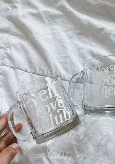 two clear mugs with white lettering on them sitting on top of a bed next to each other