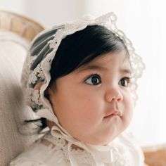 Baby girl with dark hair wearing the Penelope Lace Bonnet and matching baptism dress gazes away to the side of the camera. Fitted White Bonnet With Lace Trim, Elegant Lace Bonnet With Lace Trim, Adjustable Lace Wedding Bonnet, Wedding Bonnet With Lace Trim, Elegant Organza Lace For Ceremony, White Adjustable Lace Bonnet, White Lace Adjustable Bonnet, Elegant Adjustable Cream Bonnet, Scalloped Lace Tulle Fabric