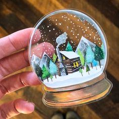 a hand holding a snow globe with a cabin in the woods painted on it's side