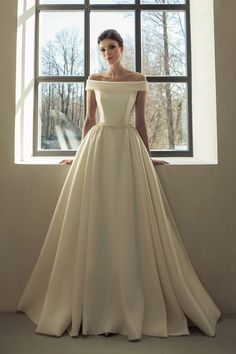 a woman standing in front of a window wearing a wedding dress with off the shoulder sleeves