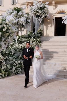 the bride and groom are walking down the steps together in front of white floral arrangements