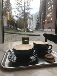 two cups of coffee and cookies on a tray in front of a window overlooking a street