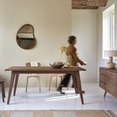 a woman walking past a wooden table in a room with white walls and flooring