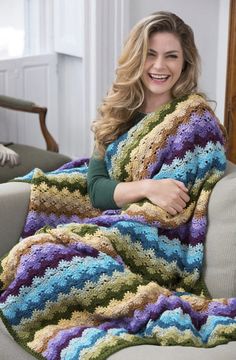 a woman sitting on a couch holding a multicolored crocheted blanket in front of her